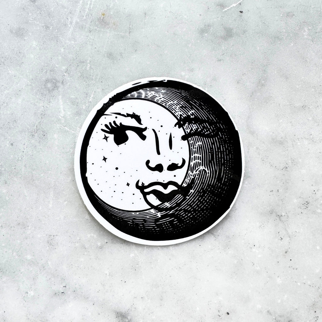 Black and white circular sticker with an illustration of a feminine face peeking out behind a crescent moon on a textured marble surface