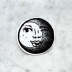 Black and white circular sticker with an illustration of a feminine face peeking out behind a crescent moon on a textured marble surface