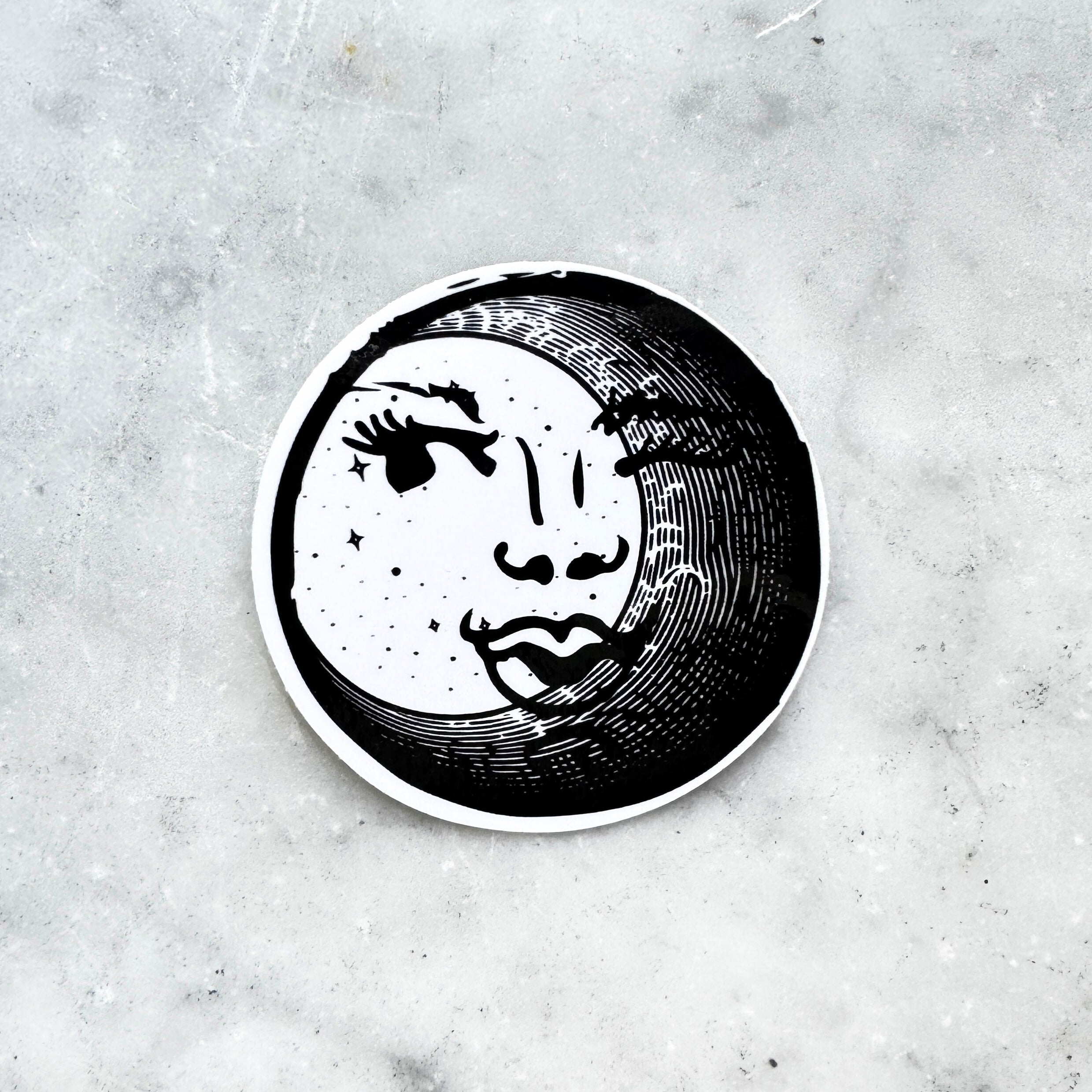 Black and white circular sticker with an illustration of a feminine face peeking out behind a crescent moon on a textured marble surface