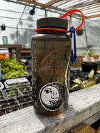 32 oz Clear water bottle with a black and white circular sticker of an illustration of a feminine face peeking out behind a crescent moon. The water bottle is sitting on a wooden ledge inside of a greenhouse.