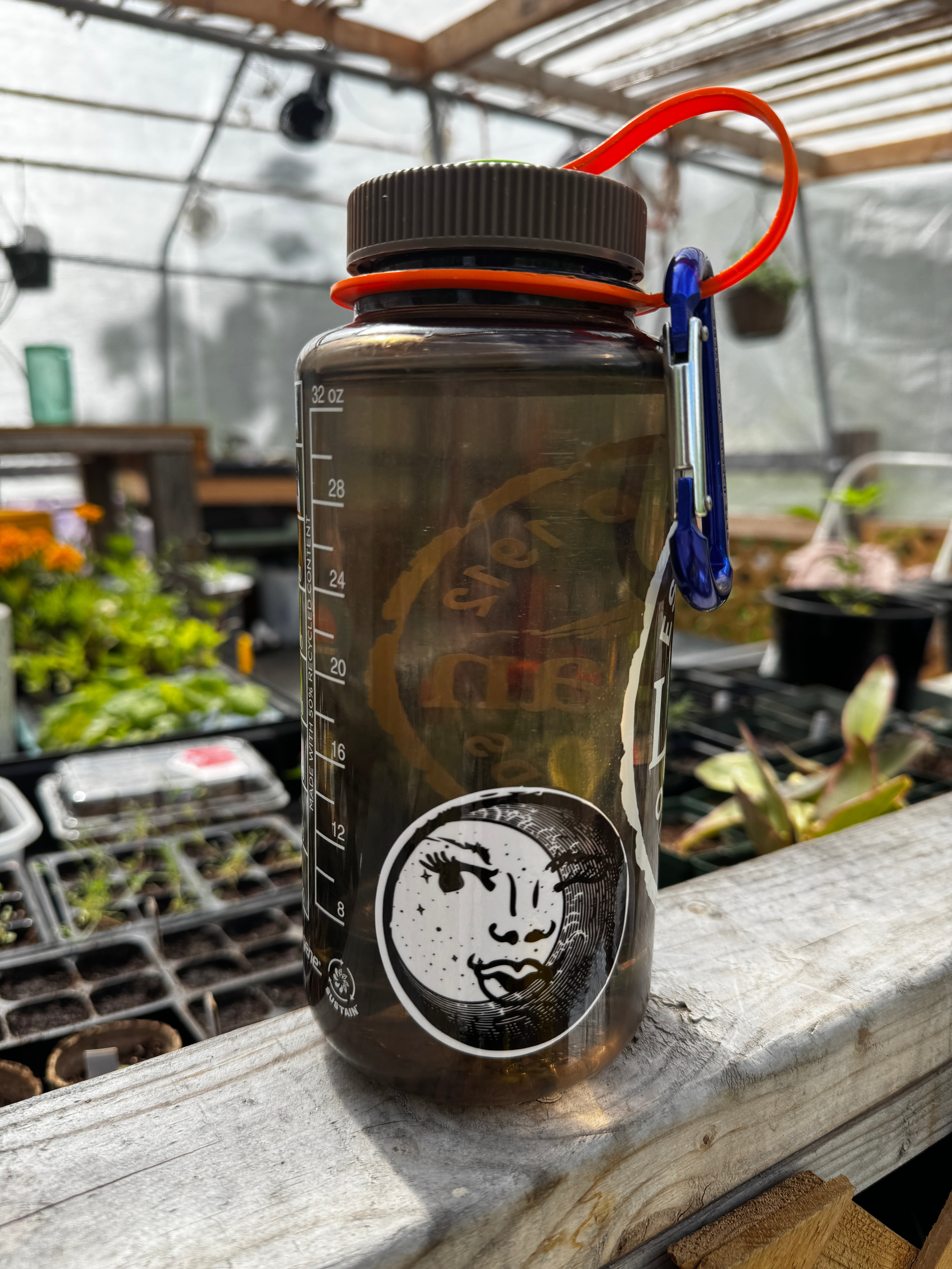 32 oz Clear water bottle with a black and white circular sticker of an illustration of a feminine face peeking out behind a crescent moon. The water bottle is sitting on a wooden ledge inside of a greenhouse.