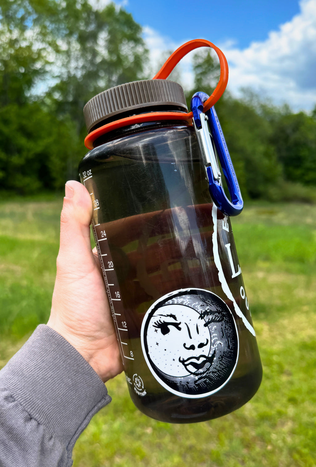 Hand holding a clear tinted water bottle that has a black and white circular sticker with an illustration of a feminine face peeking out behind a crescent moon on it. Outdoors.