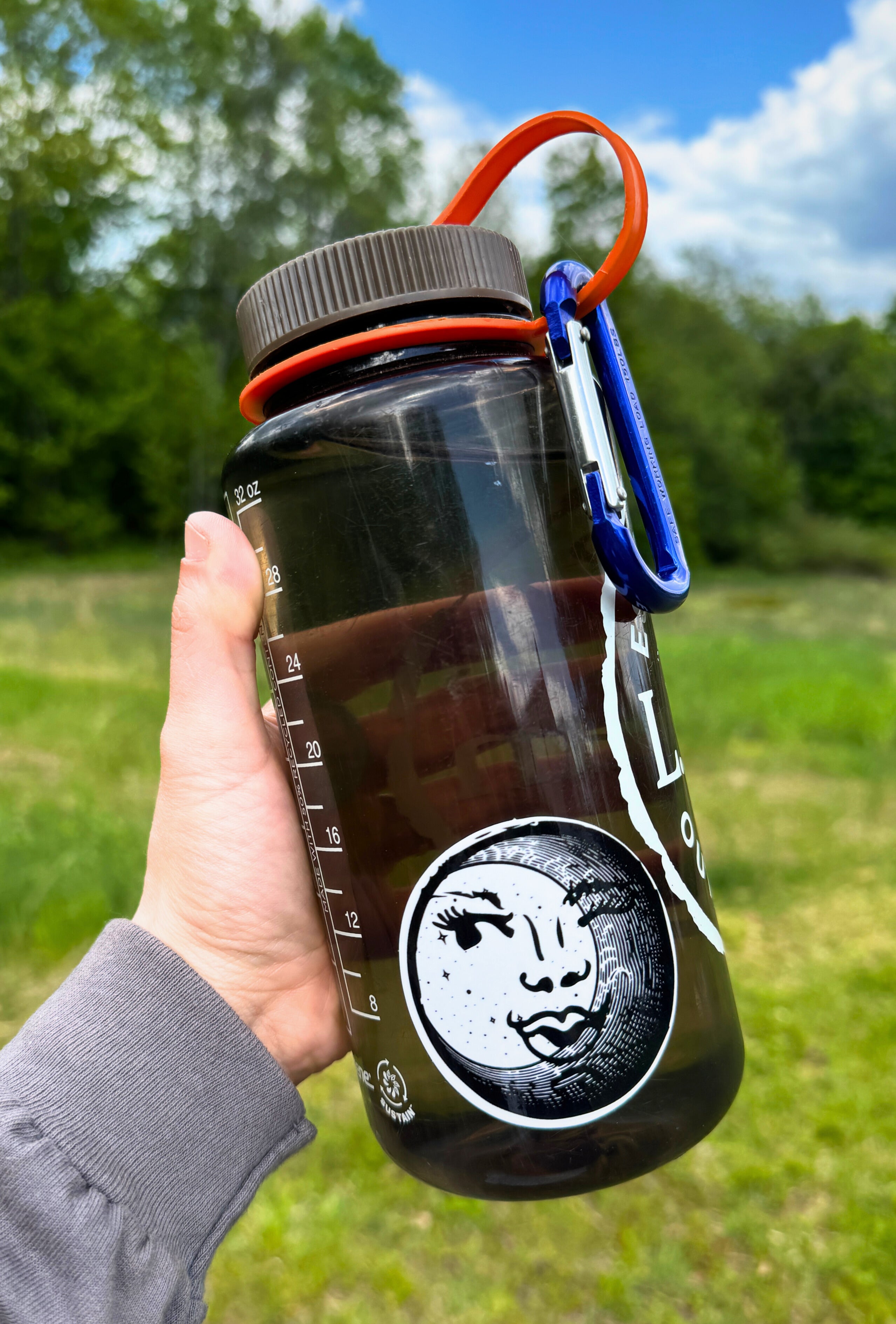 Hand holding a clear tinted water bottle that has a black and white circular sticker with an illustration of a feminine face peeking out behind a crescent moon on it. Outdoors.
