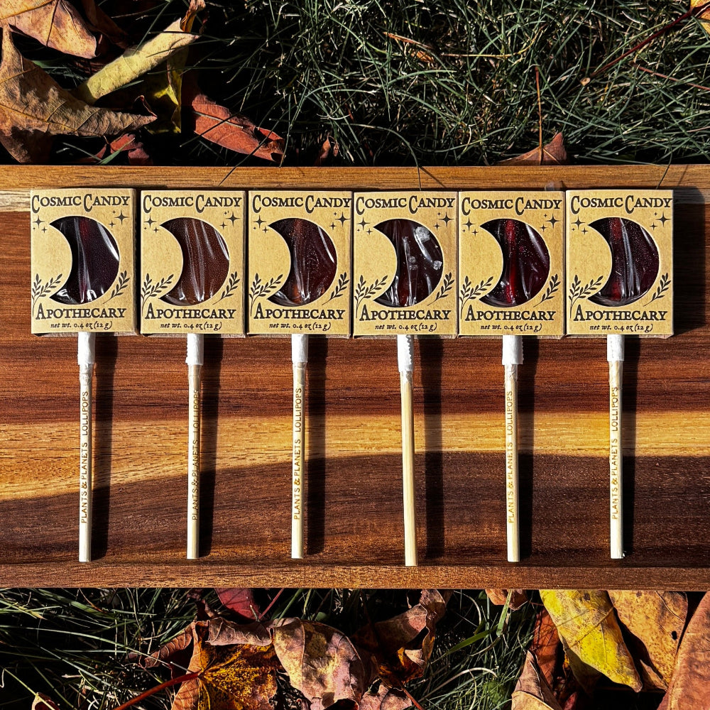 A wooden board holding six lollipops with wooden sticks encased in a kraft sleeve with a crescent moon window, arranged on grass surrounded by scattered autumn leaves. 