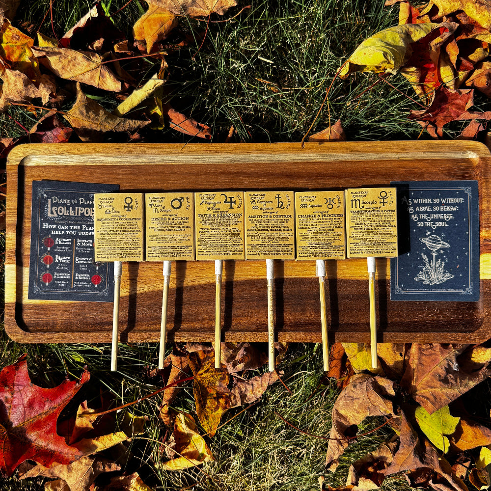 Six lollipops placed on a wooden tray with the back of the lollipop sleeve facing up showing the planetary correspondences of each flavor, displayed outdoors on a bed of fallen autumn leaves and grass.