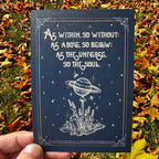 A hand holds a dark navy art print outdoors over a bed of fallen autumn leaves. The print features white celestial line art of Saturn on a lollipop stick stuck in a bed of crystals and lavender, surrounded by stars and bees, with the quote 'As within, so without; as above, so below; as the universe, so the soul.