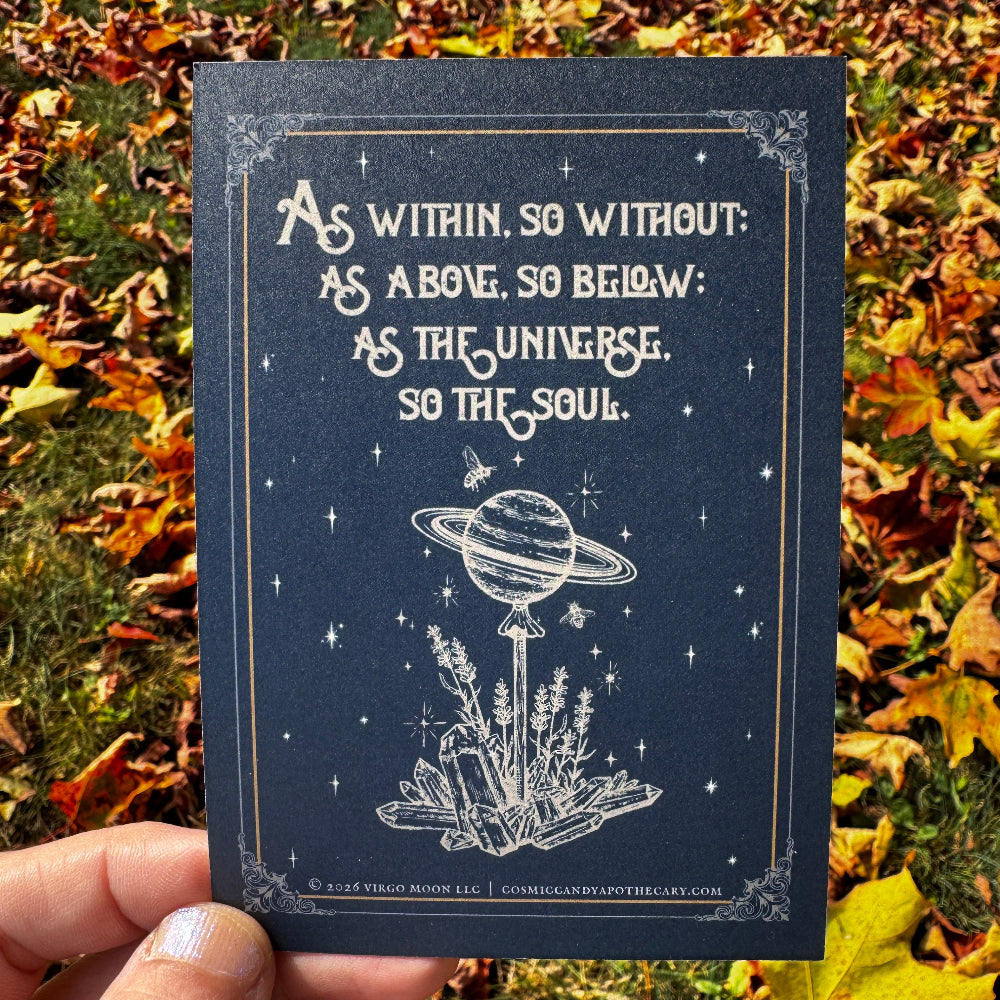 A hand holds a dark navy art print outdoors over a bed of fallen autumn leaves. The print features white celestial line art of Saturn on a lollipop stick stuck in a bed of crystals and lavender, surrounded by stars and bees, with the quote 'As within, so without; as above, so below; as the universe, so the soul.
