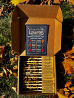 An open cardboard gift box filled with small lollipops sits outdoors on grass surrounded by fallen autumn leaves. The box lid shows a branded card reading 'Plants and Planets Lollipops: How can the Planets help you today?,' and the lollipops are arranged in rows with labeled wooden sticks visible inside.