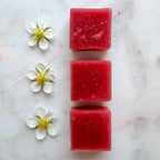 A minimalist flat lay on a white marble background with subtle grey and pink veining. Three square, semi-translucent pink fruit chews are arranged in a perfectly straight vertical line down the center of the frame, showing a fine, sugary texture and flecks of real fruit. Directly to the left of each candy sits a single, delicate white strawberry blossom with five petals and a bright yellow-green center, mirroring the vertical alignment for a clean, symmetrical composition.