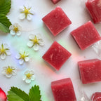 A top-down, close-up shot of several square, semi-translucent pink fruit chews showing textured flecks of real fruit. The candies are arranged diagonally across a white marble background. On the left side of the frame, seven small, white strawberry blossoms with bright yellow centers are scattered alongside vibrant green serrated leaves. A subtle, prismatic rainbow reflection casts soft hues of green, blue, and pink diagonally across the marble surface. 
