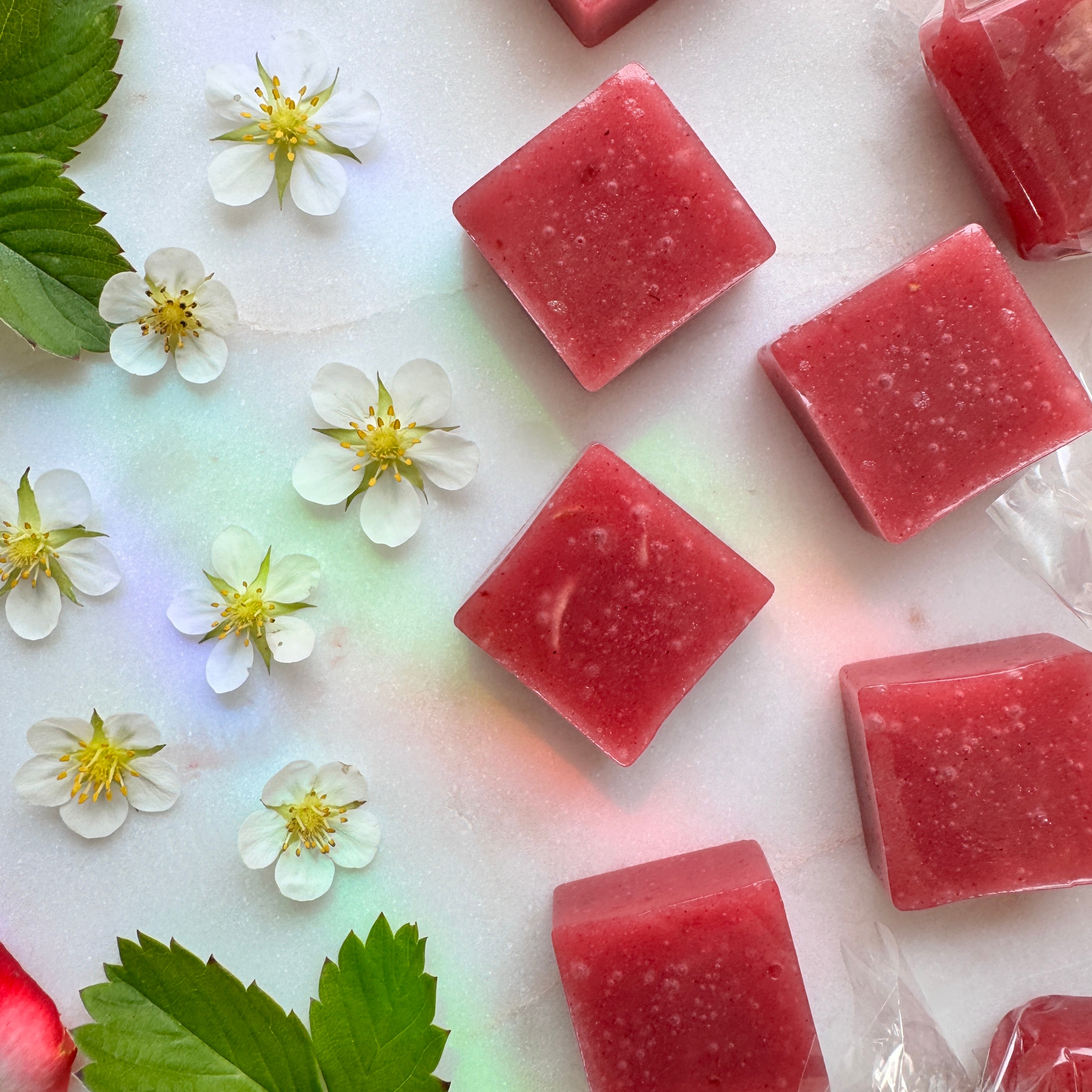 A top-down, close-up shot of several square, semi-translucent pink fruit chews showing textured flecks of real fruit. The candies are arranged diagonally across a white marble background. On the left side of the frame, seven small, white strawberry blossoms with bright yellow centers are scattered alongside vibrant green serrated leaves. A subtle, prismatic rainbow reflection casts soft hues of green, blue, and pink diagonally across the marble surface. 