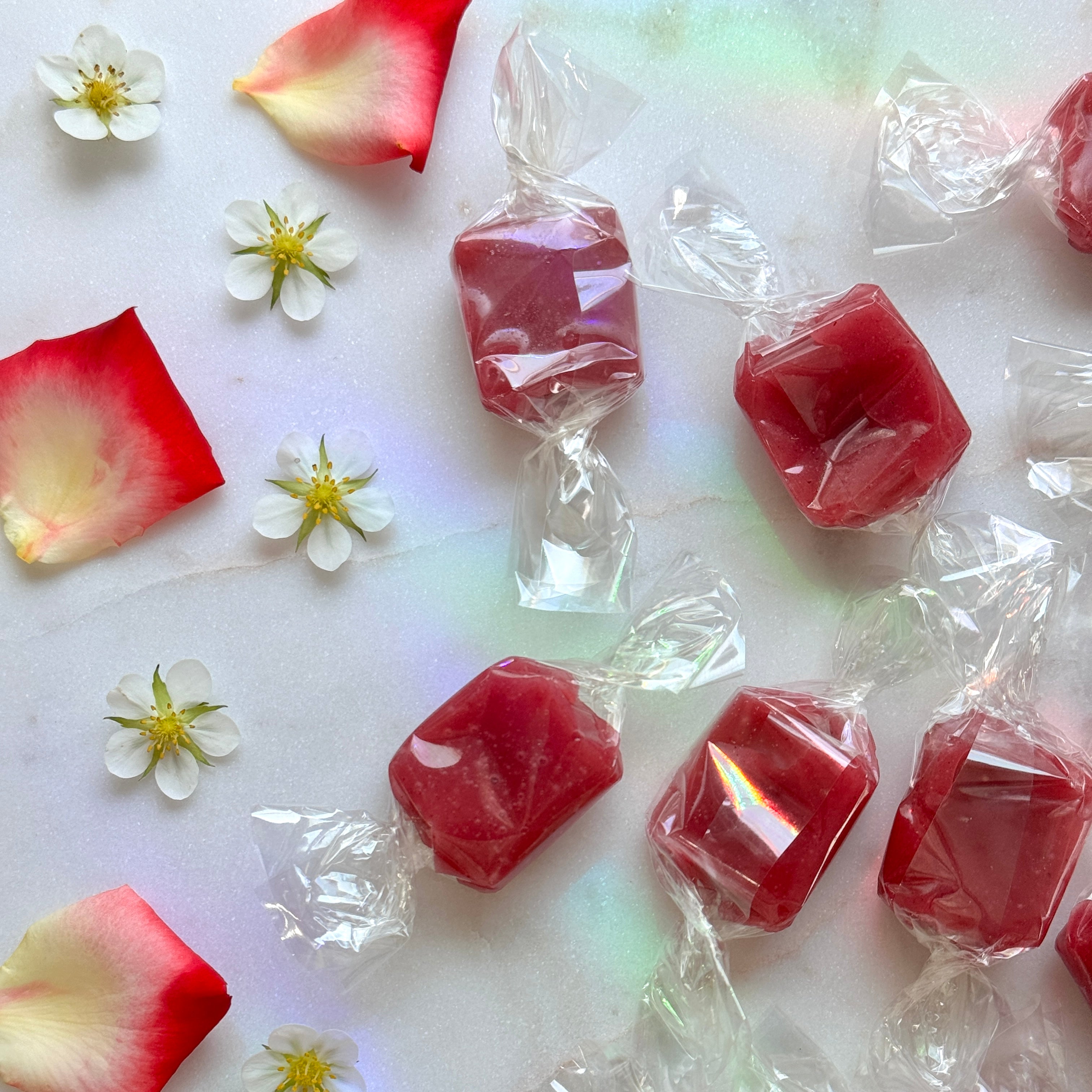 A top-down, close-up shot featuring several rectangular, strawberry-pink fruit chews individually wrapped in clear, twisted cellophane. The candies are scattered across a light white marble backdrop. On the left side of the frame, the candies are artfully balanced by four small, five-petaled white strawberry blossoms with yellow centers and three ombre rose petals that fade from deep pinkish-red to a soft yellow base. An iridescent, prismatic rainbow light leak casts a dreamy, colorful glow.