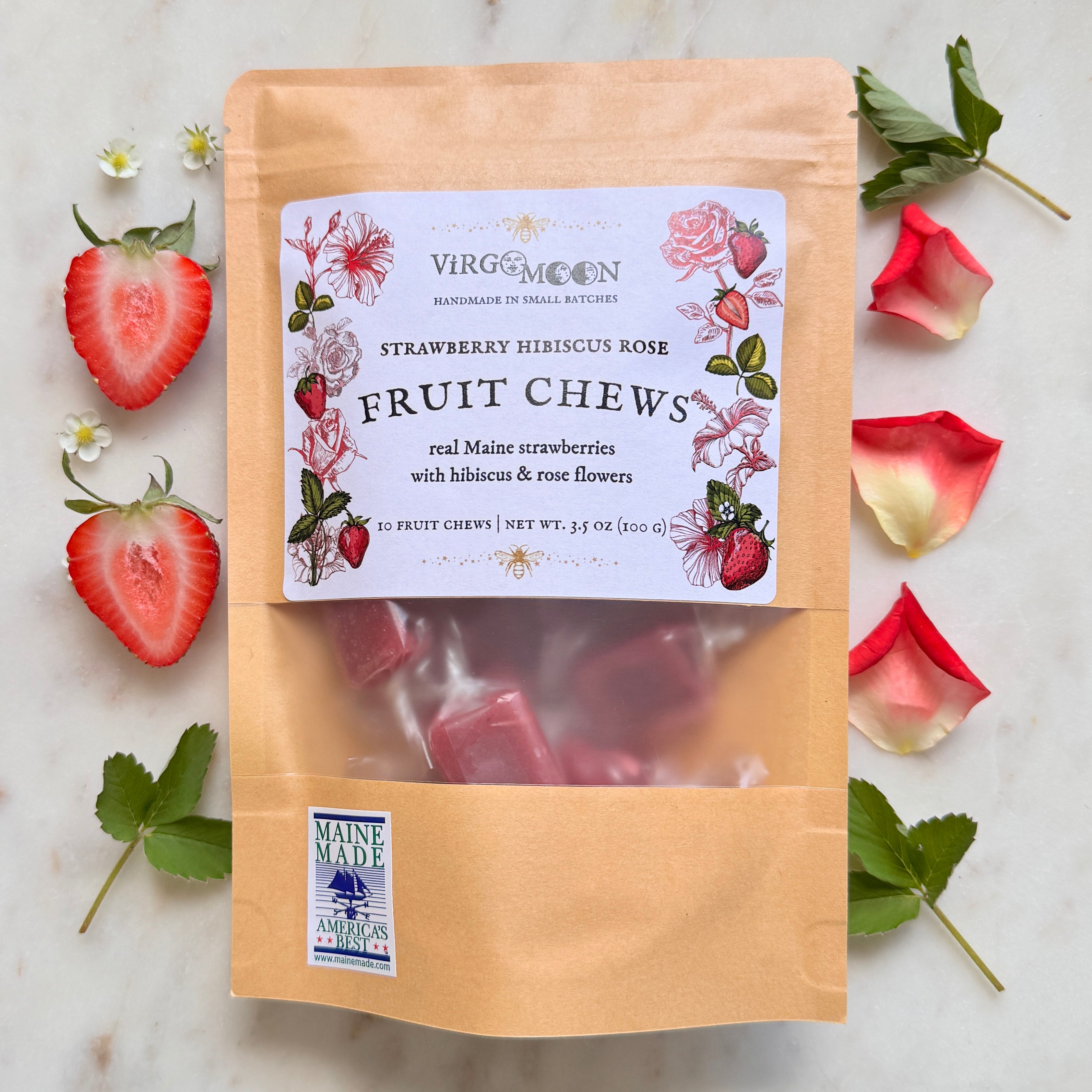 A top-down view of a Virgo Moon product package on a light marble background. The standing brown kraft pouch has a white rectangular label reading "Virgo Moon Handmade in Small Batches" with a crescent moon logo, featuring the product name "Strawberry Hibiscus Rose Fruit Chews made with real Maine strawberries with hibiscus & rose flowers." The label is framed by delicate botanical illustrations of red strawberries, hibiscus, and roses. A clear window on the pouch reveals individually wrapped chews.