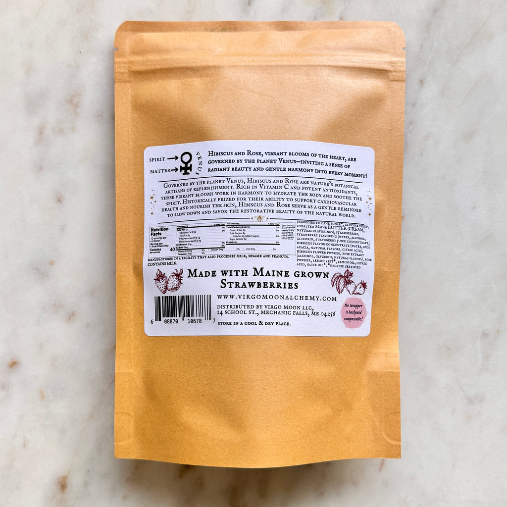 A top-down shot of the back of a brown kraft paper candy pouch centered on a white marble background. A large, rectangular white label covers the middle of the pouch.  At the top left of the label, a diagram displays the astrological symbol for Venus with arrows pointing to "Spirit" and "Matter," accompanied by text reading, "Hibiscus and Rose... are governed by the planet Venus." Below this, a paragraph details the botanical and historical benefits of Hibiscus and Rose.  