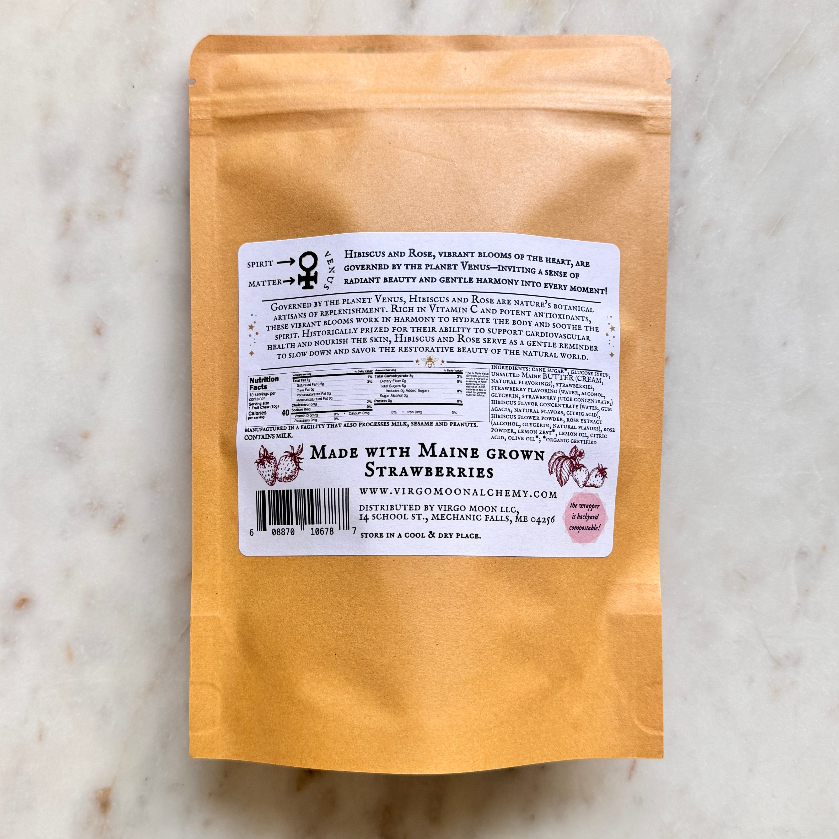A top-down shot of the back of a brown kraft paper candy pouch centered on a white marble background. A large, rectangular white label covers the middle of the pouch.  At the top left of the label, a diagram displays the astrological symbol for Venus with arrows pointing to "Spirit" and "Matter," accompanied by text reading, "Hibiscus and Rose... are governed by the planet Venus." Below this, a paragraph details the botanical and historical benefits of Hibiscus and Rose.  