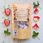 This is a flat-lay photograph of a brown kraft paper product pouch centered on a light marble surface, surrounded by fresh ingredients. The pouch label reads "Virgo Moon" and "Strawberry Hibiscus Rose Lozenges" with a description mentioning "real strawberries, hibiscus & rose flowers" and "Maine wildflower honey." A clear window shows wrapped reddish-brown lozenges inside. Two strawberry halves are on the left, rose petals and leaves are on the right, and the Maine Made logo is in the bottom-left corner.