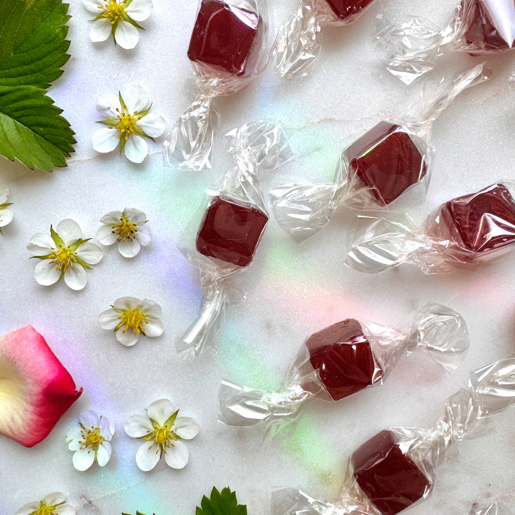 A top-down, slightly different close-up composition on the white marble surface, featuring the same individually wrapped reddish-brown lozenges scattered with white strawberry blossoms, green leaves, and a red rose petal. A different, partial fresh strawberry is visible in the bottom-left corner. The marble shows subtle iridescent reflections, similar to the previous scene but with a distinct, more spread-out arrangement.