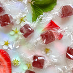 A close-up photograph capturing individually wrapped, cube-shaped reddish-purple lozenges scattered across a white marble surface. Interspersed among the clear-wrapped candies are fresh strawberry halves, delicate white blossoms with yellow centers, green serrated strawberry leaves, and red rose petals. The marble surface shows subtle iridescent light reflections under soft, natural light.
