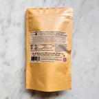A close-up photograph of the back of the brown kraft paper pouch seen in the previous image, placed on a white marble background. The white label contains text, including 'MADE WITH WILDFLOWER HONEY & STRAWBERRIES FROM MAINE,' a Nutrition Facts panel for 20 servings per container, and manufacturer information for 'VIRGO MOON LLC.' A small pink sticker near the barcode states 'this wrapper is backyard compostable!'.